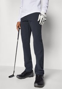Pantalons de golf navy avec une texture lisse, coupe slim et poches latérales, associés à des chaussures de sport noires et à une chemise à manches longues blanche.