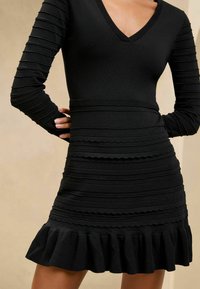 Schwarzes Langarmkleid mit V-Ausschnitt, gerippter Optik, geschichteten horizontalen Bändern und einem Rüschensaum. Hergestellt aus einem dehnbaren Material.