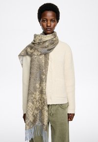 Cárdigan de lana en color crema con patrón texturizado, combinado con una bufanda estampada que presenta tonos dorados y grises, terminada con flecos en azul claro.