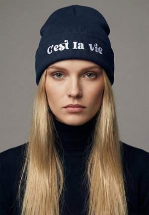 Navy ribbed knit beanie with embroidered text "C'est la Vie" in white. Soft texture, snug fit, unisex design. Suitable for cold weather.