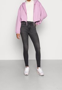 Sudadera corta de color púrpura claro con cordón, cuello alto de canalé blanco, jeans ajustados grises y zapatillas blancas con acentos rosas.