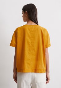 Korte mouwen, losse top van gestructureerde, mosterdgele stof met een ronde hals en een rechte zoom, gecombineerd met lichte broek.