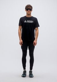 Μαύρο μπλουζάκι με λευκό κείμενο, μαύρο σορτς, μαύρα κολάν και παπούτσια με πράσινες λεπτομέρειες. Το μοντέλο φοράει σκούρα γυαλιά ηλίου, στέκεται μπροστά σε ένα απλό φόντο.