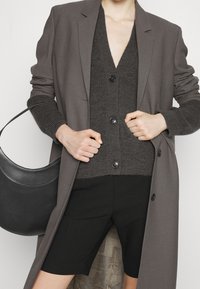 Lätt grå blazer över en mörkgrå stickad cardigan med knappar, parat med svarta shorts. En böjd svart väska hänger över armen.