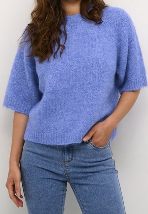 Kobieta w miękkim, jasnofioletowym sweterku z krótkim rękawem, zestawionym z wysokim stanem niebieskich dżinsów, z jedną ręką w kieszeni.