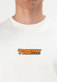 Close-up of a person wearing a white shirt with "TOMmy jeans" embroidered in bold orange and black letters on the chest.