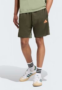 Pantaloncini sportivi verde oliva con fascia elastica in vita e un logo arancione come dettaglio. Abbinati a calzini a righe e sneaker bianche con strisce verdi.