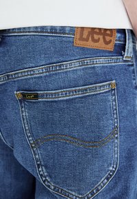 Modri jeans iz denima z rjavim usnjenim napisom z logotipom "Lee" na pasu, z zlatimi šivi in detajli na zadnjem žepu.