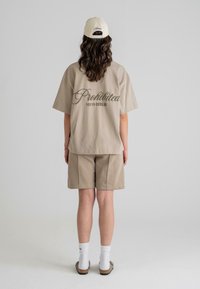 Beige pamut póló, hátul sötétzöld "Prohibited 10119 Berlin" felirattal, hozzáillő sorttal és világos sapkával párosítva.