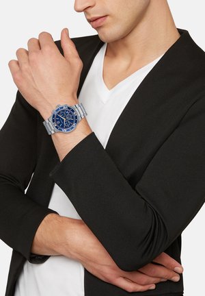 Blå chronograph ur med sølvarmbånd, placeret på et håndled. Uret har en tekstureret blå skive og fed hvide tal.
