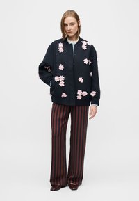 Geacă bomber bleumarin cu aplicații florale roz, cu decolteu rotund și închidere cu nasturi în față. Asortată cu pantalon larg cu dungi închise la culoare.