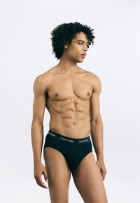 Čierne bavlnené slipy s elastickým pásom, ktorý obsahuje biele logo "Calvin Klein". Model stojí s neutrálnym výrazom.