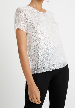 Nainen, jolla on lyhythihainen valkoinen paita, jossa on hopeisia paljetteja, yhdistettynä istuviin mustiin housuihin yksinkertaisen taustan edessä.