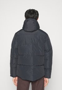 Doudoune marine avec capuche, présentant des sections matelassées horizontales, un ourlet droit et une texture lisse et brillante.