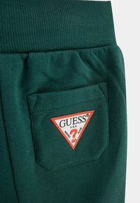 Žalias tekstūruotas audinys su šonine kišene, kurioje yra trikampis raudonas ir baltas "GUESS" logotipas. Matoma raukšlėta juostos detalė.