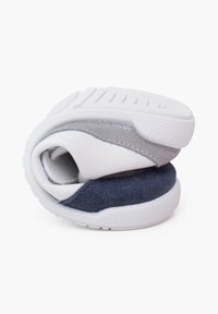 Baskets avec une tige synthétique blanche, des accents en suede gris et marine, et une semelle en caoutchouc texturé. Position roulée montrant la flexibilité et le design.