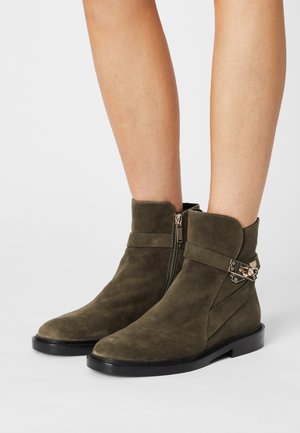 Enkellaarsjes van olijfgroen suede tot aan de enkel met een rits aan de zijkant, decoratieve gouden gesp en een platte zwarte zool, gedragen op blote benen.