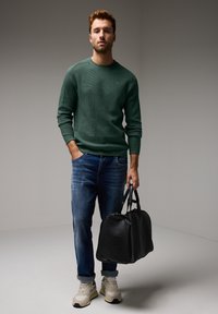 Zelený texturovaný pletený svetr, modré džíny, béžové tenisky, držící černou koženou cestovní tašku. Neutrální pozadí, neformální vzhled.