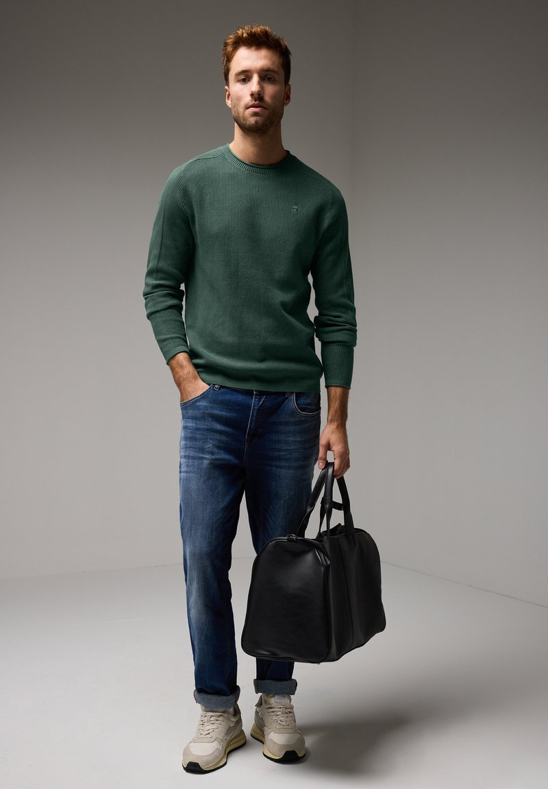 Zelený texturovaný pletený svetr, modré džíny, béžové tenisky, držící černou koženou cestovní tašku. Neutrální pozadí, neformální vzhled.