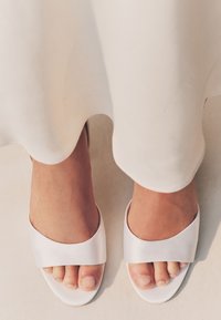 Piedi che indossano sandali bianchi aperti con una larga fascia, parzialmente coperti da un indumento bianco fluente su un pavimento chiaro.