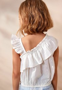 Biała bezrękawnikowa bluzka z falbaniastymi ramiączkami, teksturowanym materiałem w grochy oraz elastycznym paskiem w talii. Widok z tyłu, prezentujący wzór.