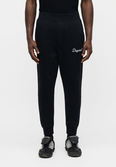 Μαύρη φόρμα με λευκό λογότυπο "Dsquared2" στο αριστερό μηρό, φορεμένη με μαύρα και λευκά αθλητικά παπούτσια Dsquared2, απεικονιζόμενη σε άτομο που στέκεται σε λευκό φόντο.