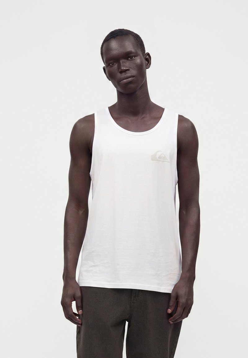 Jeune homme portant un débardeur blanc uni sans manches et un pantalon foncé, debout avec une expression neutre devant un fond uni.