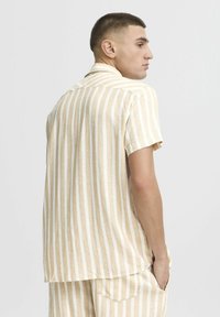 Camisa de manga corta en beige claro con rayas verticales blancas. Tejido suave, corte relajado, diseño de cuello. La vista trasera resalta la costura y la textura.
