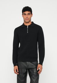 Suéter preto de malha com gola com fecho, design ajustado e punhos canelados, combinado com calças de pele pretas. Aparência minimalista e texturada.