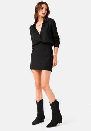 Woman wearing a black long-sleeve blouse, black mini skirt, and black mid-calf boots standing against a plain light background.