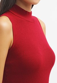 Vrouw in een mouwloze, hoge hals, geribbelde rode top, weergegeven van schouders tot het midden van de torso tegen een effen witte achtergrond.