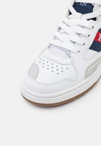 Detailní záběr na bílé sportovní tenisky s bílými šňůrami, perforovanou špičkou, námořnickou a červenou logovou nášivkou a béžovou texturovanou podrážkou.