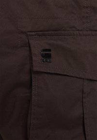 Material textil de culoare maro cu o textură netedă, având o buzunar vizibil și un patch cu logo-ul "G-RAW" de culoare neagră cusut pe lateral.