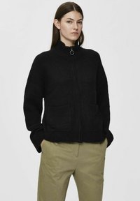Svart zip-up jakke laget av mykt, teksturert stoff med høy krage, to frontlommer og avslappet passform, kombinert med lyse bukser.