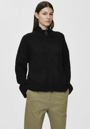 Black zip-up jacket made of soft, textured fabric with a high collar, two front pockets, and relaxed fit, paired with light-colored pants.