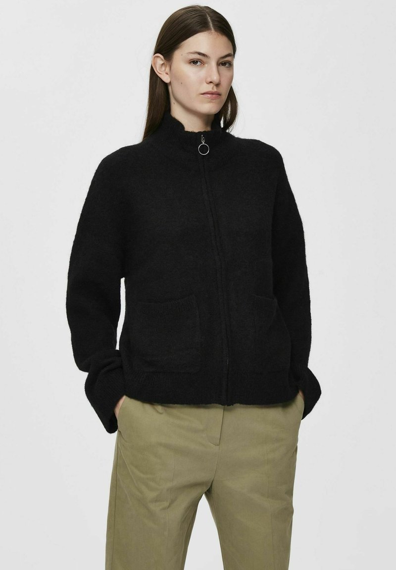 Svart zip-up jakke laget av mykt, teksturert stoff med høy krage, to frontlommer og avslappet passform, kombinert med lyse bukser.