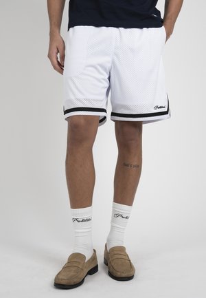 Męskie nogi w białych siateczkowych szortach z czarnym obszyciem, białe skarpetki z napisem „Prohibitions” oraz jasnobrązowe mokasyny na neutralnym tle.