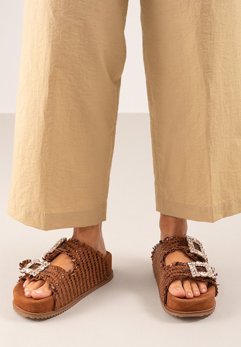 Sandalias trenzadas marrones con una suela gruesa, adornadas con dos grandes hebillas cuadradas y bordes rizados, llevadas con pantalones de pierna ancha beige.