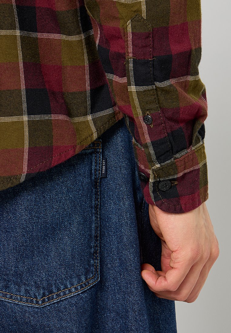 Chemise en flanelle à motif à carreaux rouge, vert et noir avec une manche à bouton, assortie à un jean bleu, mettant en valeur des textures contrastées.