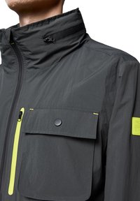 Ciemnoszara kurtka z wysokim kołnierzem, z kieszenią z zapięciem na nap i jasnółożowym zamkiem błyskawicznym, wykonana z lekkiego materiału.