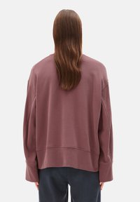 Person mit langen braunen Haaren, die einen lockeren mauvefarbenen Pullover und dunkle Hosen trägt, steht mit dem Rücken zur Kamera vor einem schlichten Hintergrund.