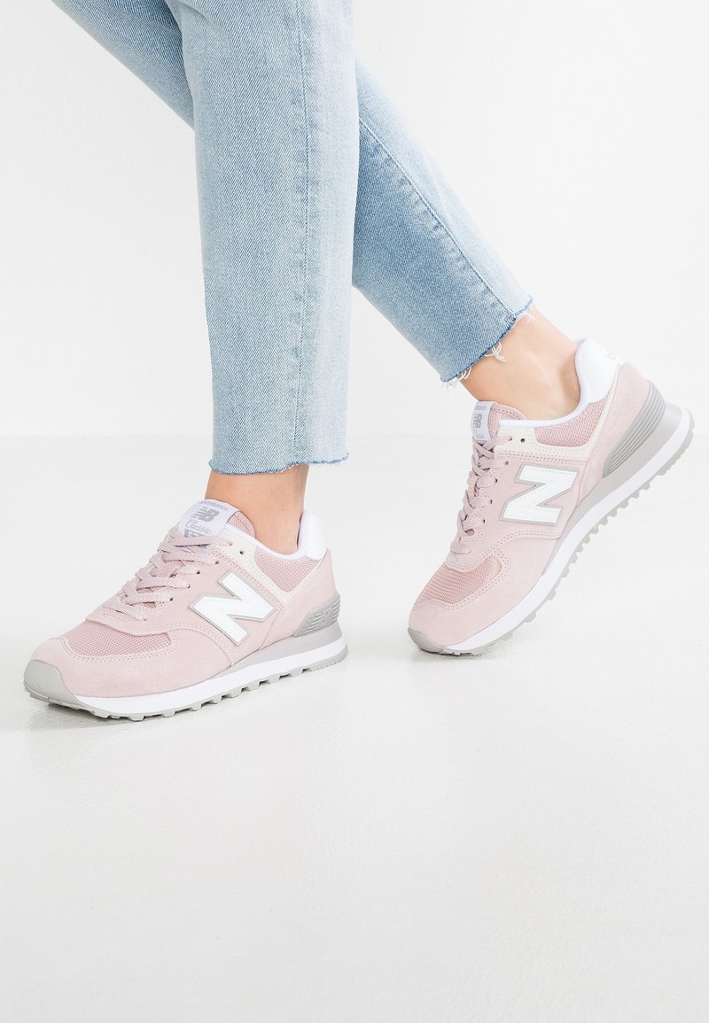 Personne portant un jean bleu clair et des baskets New Balance roses avec des accents blancs, debout sur un sol blanc devant un fond uni.