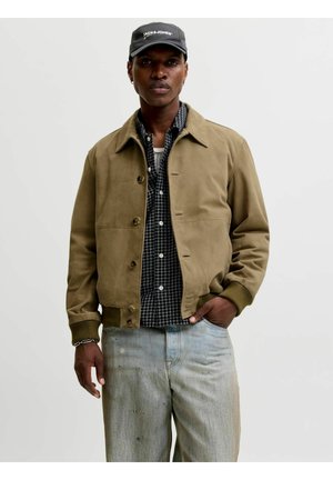 Person trägt eine beige, geknöpfte Jacke, ein schwarz-weiß kariertes Hemd, hellblaue Jeans und eine schwarze Kappe mit weißem Text.