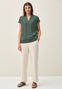Blusa verde a maniche corte con scollatura a V, abbinata a pantaloni larghi color off-white e sandali scivolati verde scuro, con una texture liscia.