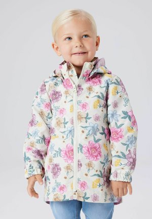 Fiatal gyermek szőke hajjal, fehér kabátban, amelyet rózsaszín, sárga, lila és kék virágminták díszítenek, egy egyszerű háttér előtt áll.