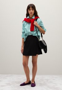 Camicia a righe verde e bianca, maglione rosso legato sulle spalle, gonna nera avvolgente, scarpe piatte viola e una borsa nera con frange.
