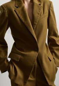 Blazer marrón a medida con solapas de muesca, cierre de un solo botón y dos bolsillos frontales. La textura de la tela parece suave y elegante.