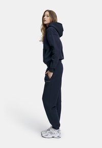 Mørkeblå hettegenser og matchende joggebukser laget av mykt stoff; har en lomme foran, mansjetter på ermene, og logo detaljer på buksene.