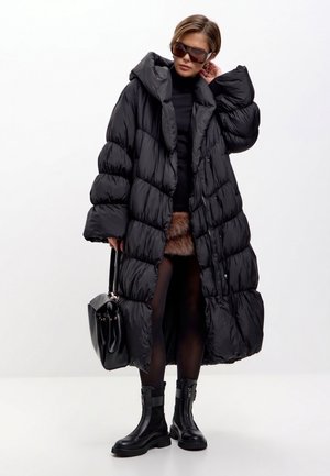 Mujer con abrigo acolchado negro de gran tamaño, botas negras y gafas de sol, sosteniendo un bolso negro sobre un fondo blanco liso.