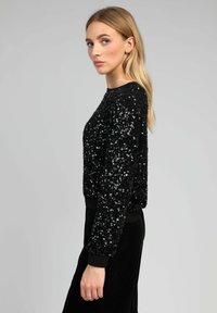 Pull en sequins noirs avec un design cropped, un col rond et des poignets côtelés. Les sequins créent une surface texturée et réfléchissante.